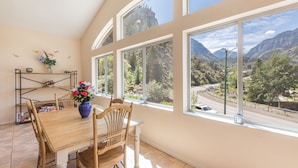 Un comedor con mesa y sillas de madera, un jarrón con flores y ventanas amplias con vistas a la montaña.