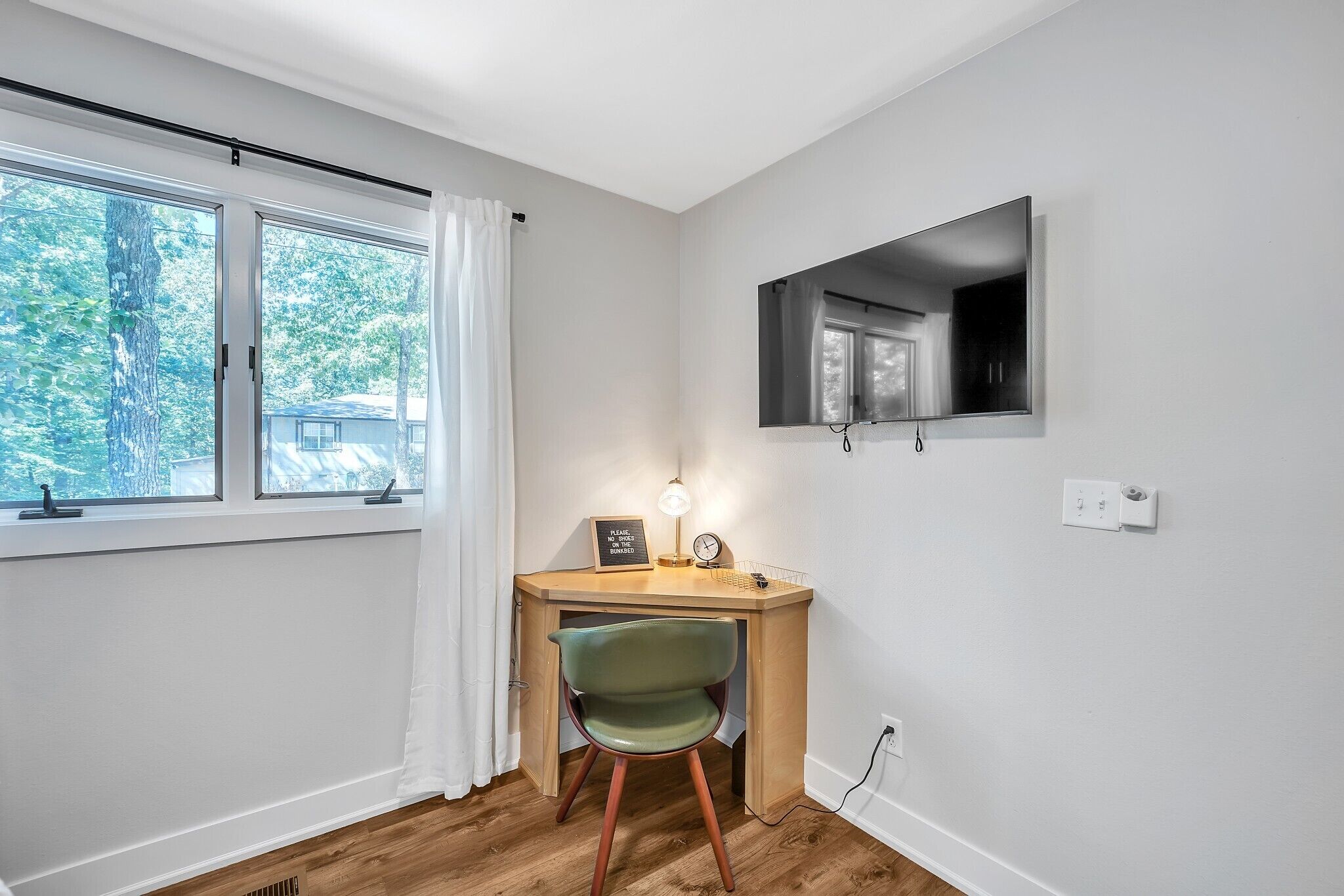 A room with a wooden desk, a green chair, a window with white curtains, and a mounted TV.