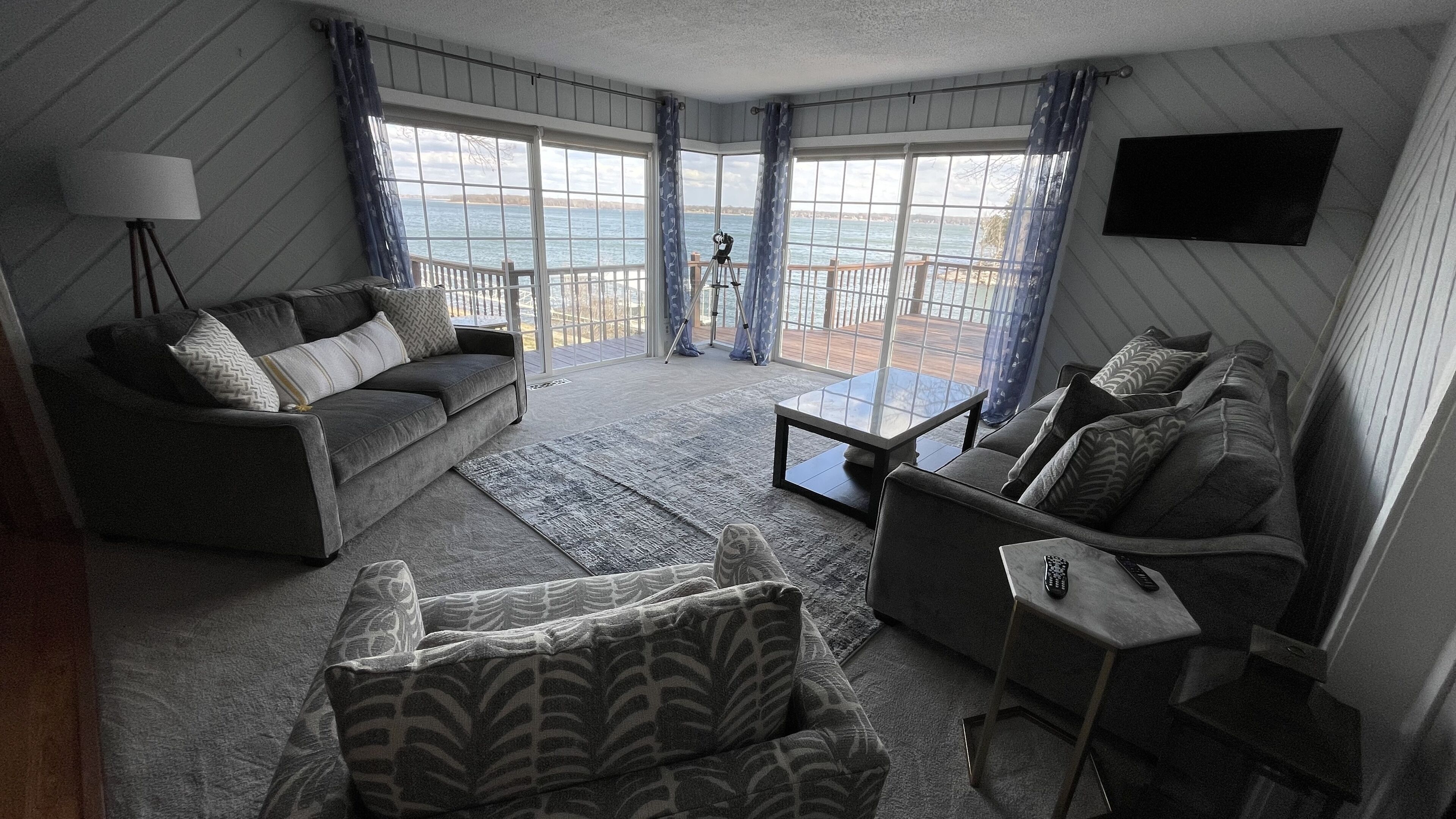 A living room with a sofa, a coffee table, and a TV mounted on the wall. There is a large window with curtains, and a balcony with a telescope.