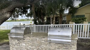 Una parrilla de piedra al aire libre con dos asadores, frente a un fondo de valla blanca, palmeras y una masa de agua.