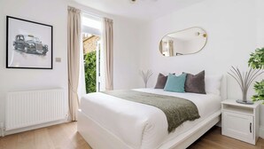 A bedroom with a white bed, a radiator, a framed picture, a mirror, and a window with curtains. - Be More Elizabeth! (Greater London)