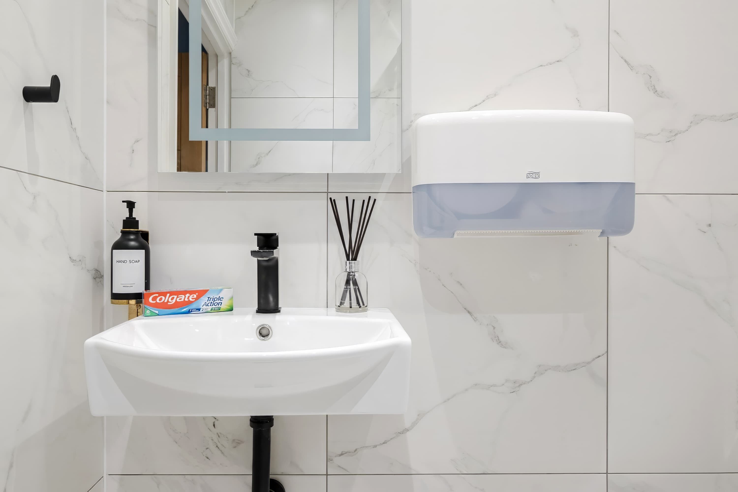 A bathroom with a white sink, a hand soap dispenser, a Colgate toothpaste box, a black soap pump, and a hand dryer.