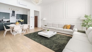 Una sala moderna con un sofá blanco, una zona de comedor con una mesa y sillas blancas, y una cocina al fondo.