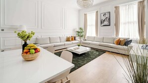 A modern living room with a white sofa set, a dining table with a fruit bowl, a coffee table, and a chandelier. - Be More Elizabeth! (Greater London)