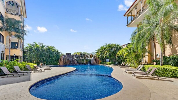 A swimming pool with lounge chairs and palm trees, surrounded by buildings. - SR9: Luxury Condo Across the Street from Beach (Tamarindo)