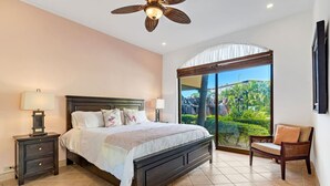 A bedroom with a large bed, a nightstand, a chair, a ceiling fan, and a view of a garden through a sliding glass door. - SR9: Luxury Condo Across the Street from Beach (Tamarindo)