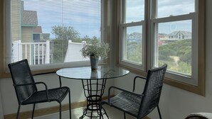Ein Zimmer mit einem runden Glastisch, zwei schwarzen Stühlen, einer Topfpflanze und großen Fenstern mit Blick auf Gebäude und Bäume.