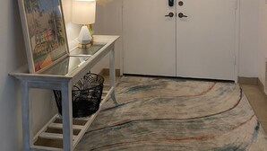 Un pasillo con una puerta blanca, una consola con espejo, un cuadro enmarco, una lámpara y una alfombra estampada.
