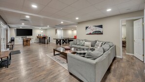 Un salon spacieux avec un canapé gris, une table basse en bois et un téléviseur à écran plat. On aperçoit à l’arrière-plan un coin repas avec une table et des chaises.