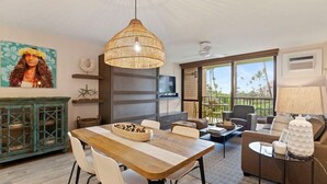Un amplio salón con una mesa de comedor de madera, sillas blancas, un sofá y un gran ventanal con vistas a palmeras.
