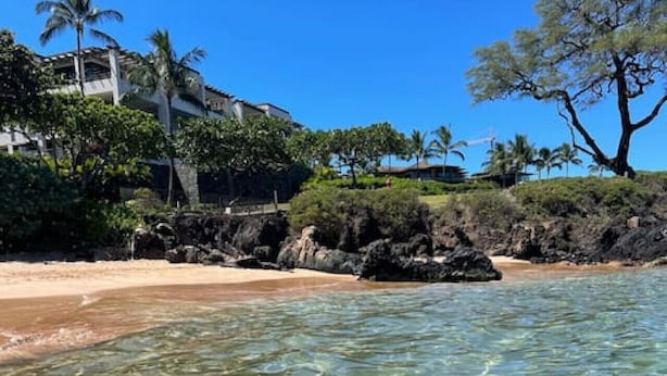 Una playa con aguas transparentes y poco profundas, una orilla arenosa y un edificio con balcón con vistas a la playa.