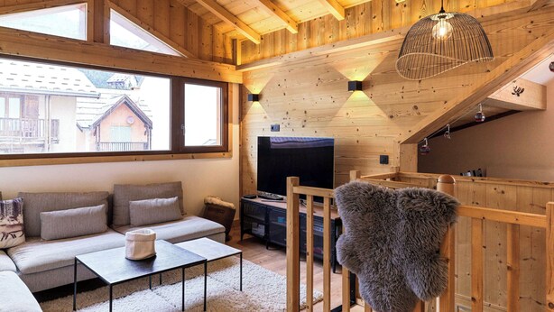 A cozy wooden cabin interior with a sofa, TV, and a view of a mountain house through large windows. - Le Chenaillet, duplex at the foot of the slopes and spa (Montgenèvre)