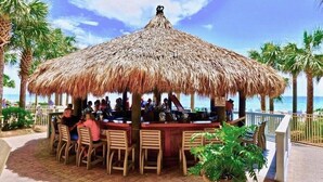 Een openluchthapbar met rieten dak, houten stoelen en tafels, omgeven door palmbomen en uitzicht op zee.