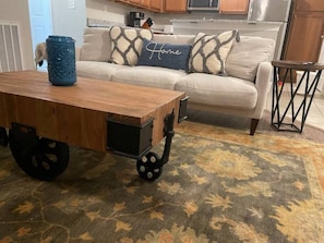 A wooden cart with wheels in a living room with a patterned rug, a sofa with decorative pillows, and a kitchen in the background.
