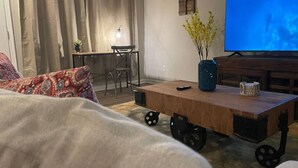 Ein Wohnzimmer mit einer Couch, einem rollbaren Couchtisch, einem Fernseher und einem kleinen Tisch mit Lampe und Pflanze.