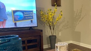 Ein Wohnzimmer mit Fernseher, der ein Schild für den Sunset Cliffs Natural Park zeigt, einer Topfpflanze und einem dekorativen Gegenstand auf Holzboden.