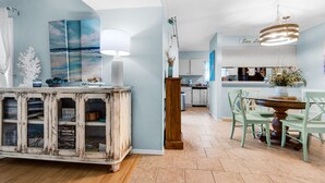 A dining area with a table and chairs, a cabinet with glass doors, and a wall with a painting. -  🏝️Short Walk 2 Gulf and Bay-Pool-Large Balcony-Boardwalk B2 (Pensacola Beach)