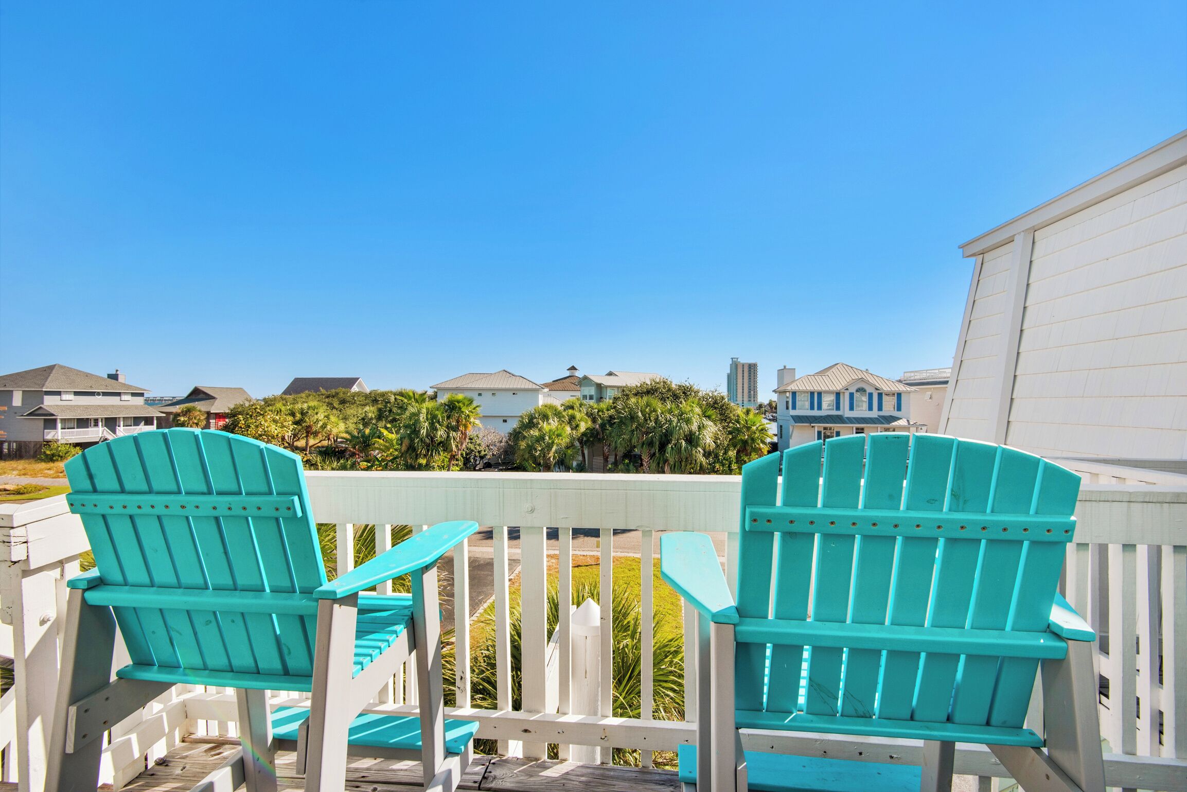 A wooden deck with two turquoise Adirondack chairs facing a white fence and a row of houses in the background.