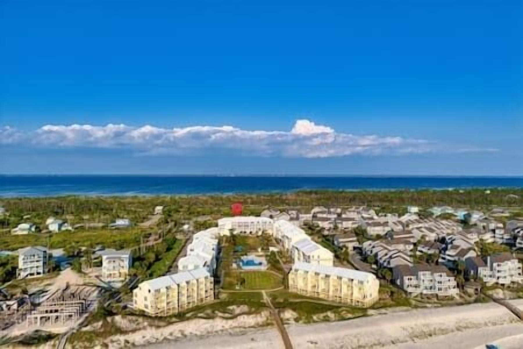 An aerial view of a coastal residential area with a central cluster of buildings, a beach in the background, and a clear sky.