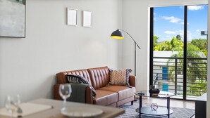 A modern living room with a brown leather sofa, a glass coffee table, a floor lamp, and a view of greenery through large windows.