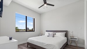 A bedroom with a large bed, a ceiling fan, a nightstand, a bedside table, and a window with a view of the city.
