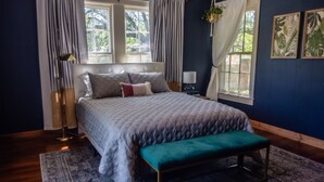 A bedroom with a large bed, a bench, a floor lamp, and a window with curtains.