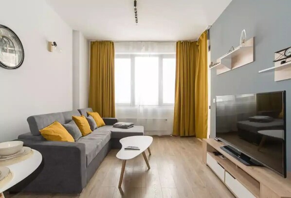 Un salon moderne avec un canapé gris, des coussins jaunes, une table basse blanche et un téléviseur à écran plat posé sur un meuble en bois.