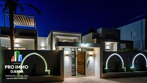 A modern house with a gated entrance, illuminated by wall-mounted lights, and a palm tree in the foreground.