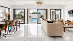 A spacious living room with a large sofa, a dining area with a table and chairs, and a view of a swimming pool through large sliding glass doors.