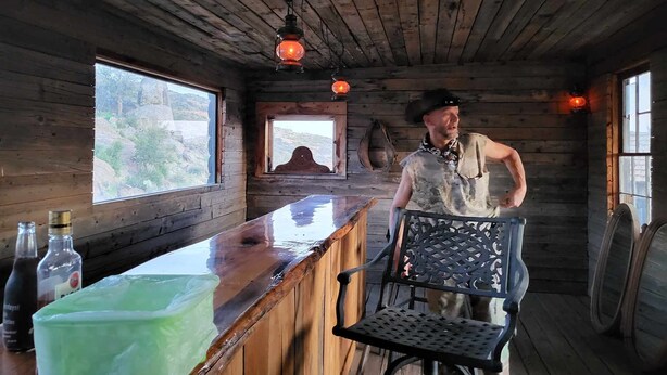 Interno di una rustica baita in legno con angolo bar, pareti in legno e una finestra che si affaccia su un paesaggio roccioso.