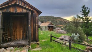 Una baita di legno rustica con un'ampia porta aperta e una piccola struttura ad essa collegata, immersa in un'area erbosa con rocce e vegetazione rada.