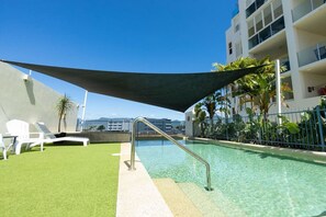 A rooftop pool area with a clear view of the city skyline, a shaded lounging area, and a metal pool ladder.