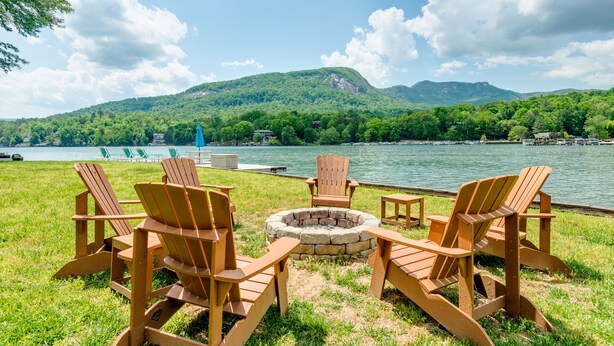 A lakeside setting with Adirondack chairs and a fire pit, surrounded by lush greenery and mountains.