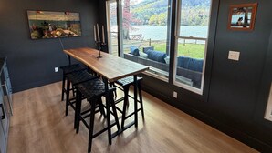 A modern kitchen with a wooden table and bar stools, large windows with a view of a lake and hills, and a painting on the wall.