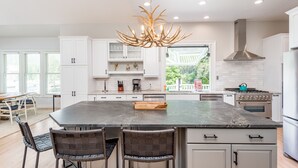 A modern kitchen with white cabinetry, a large island with bar stools, a built-in stove, and a unique antler chandelier.