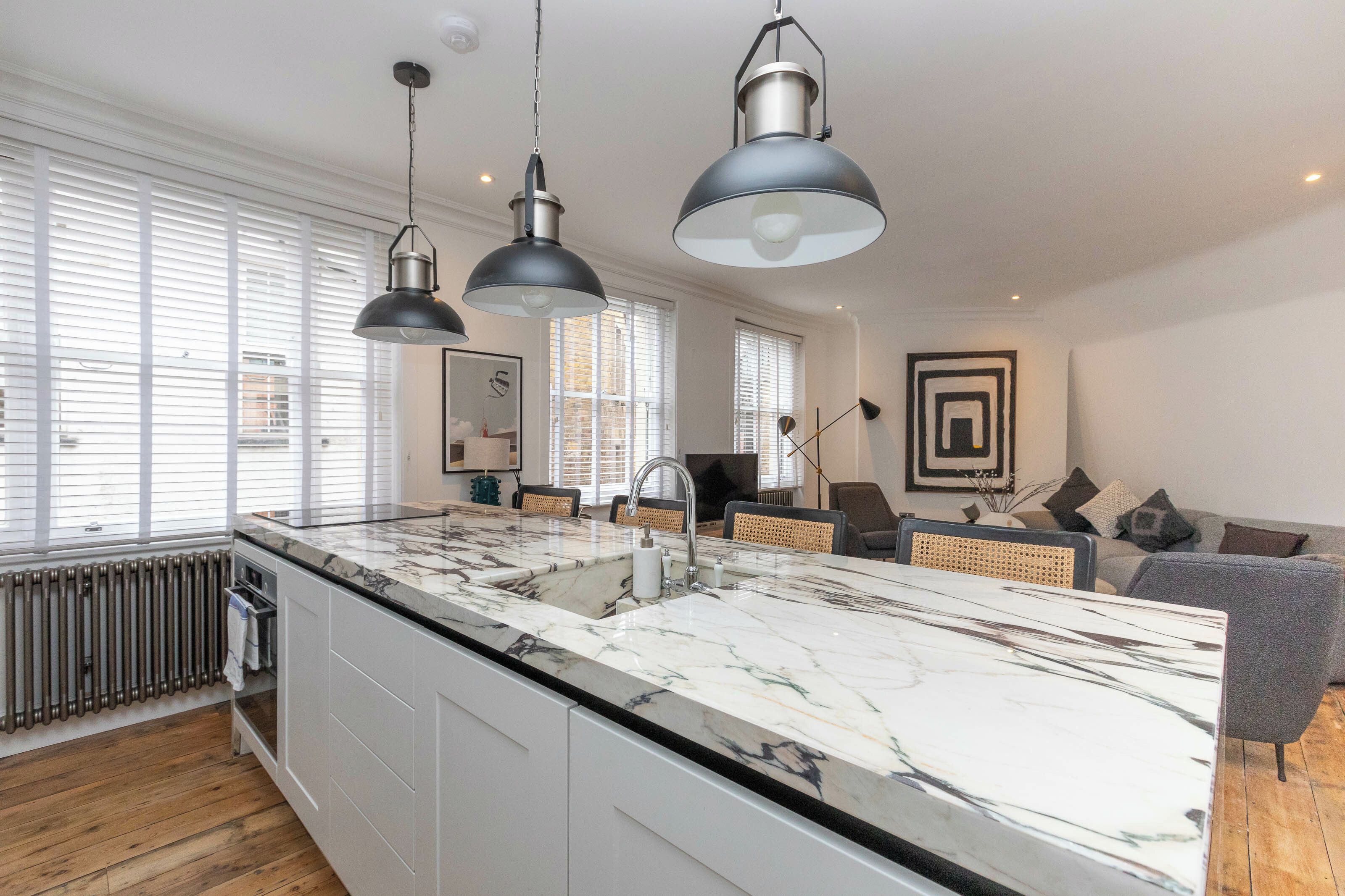 Una cocina moderna con encimera de mármol, luces colgantes y vistas al salón, que incluye un sofá y un cuadro.
