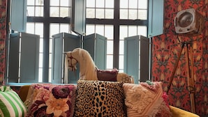 Ein Zimmer mit einem gelben Sofa, Kissen mit Blumen- und Leopardenmuster, einer Dekoration mit einem Pferdekopf und einem Wandventilator.