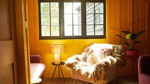 Ein gemütliches Zimmer mit einem großen Fenster, einem flauschigen Sessel, einem kleinen Tisch mit Lampe und einer Topfpflanze.
