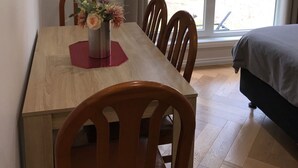A room with a wooden dining table, four chairs, a vase with flowers, a bed with a dark cover, and a sliding glass door leading to an outdoor area with trees. - Cosy apartment close to Puffing Billy  (Clematis)