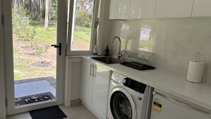 A compact kitchen with a washing machine, a sink, and a window overlooking a garden. - Cosy apartment close to Puffing Billy  (Clematis)