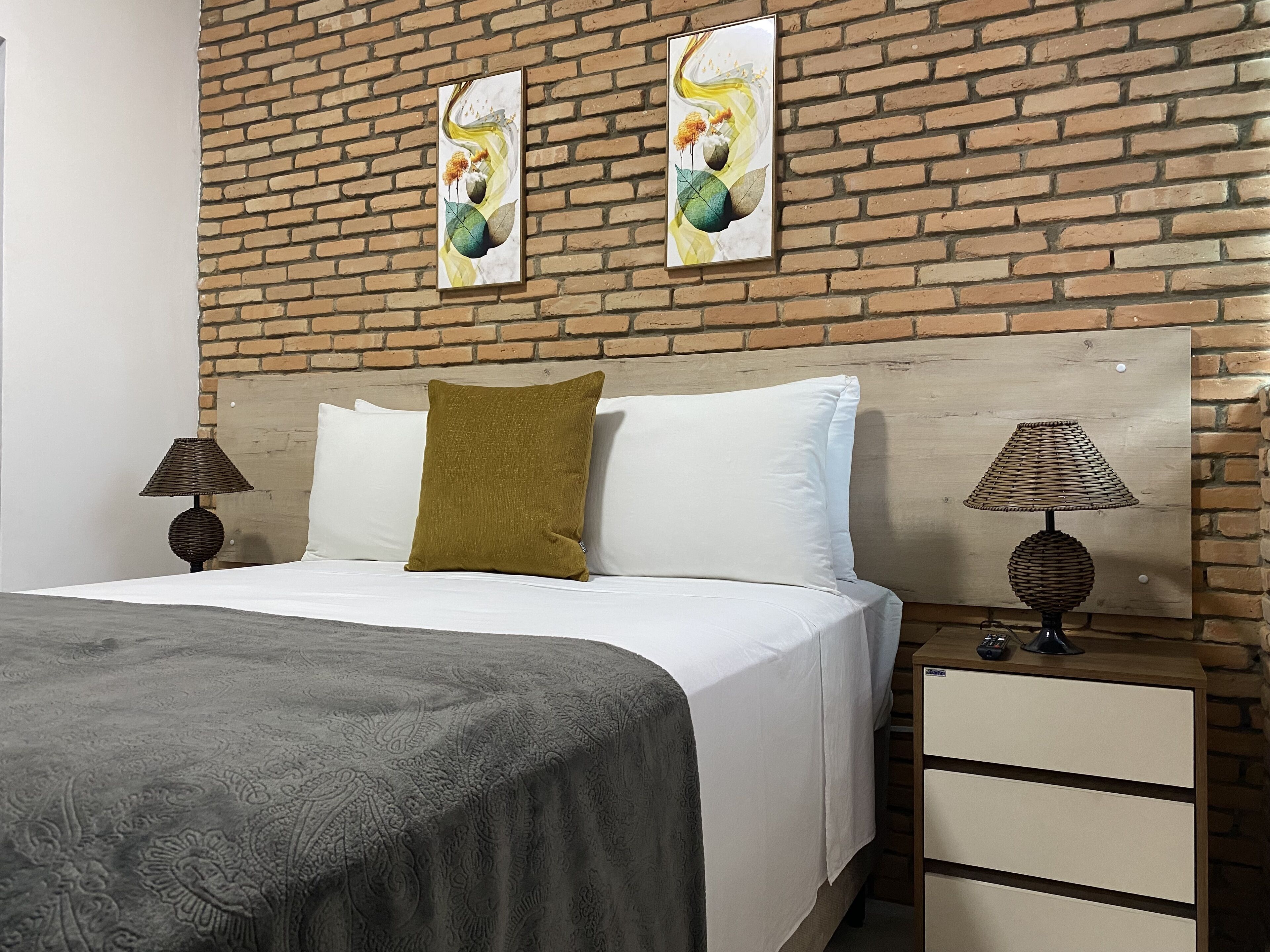 A bedroom with a brick wall, a bed with white and olive green bedding, two bedside lamps, and a wooden nightstand.