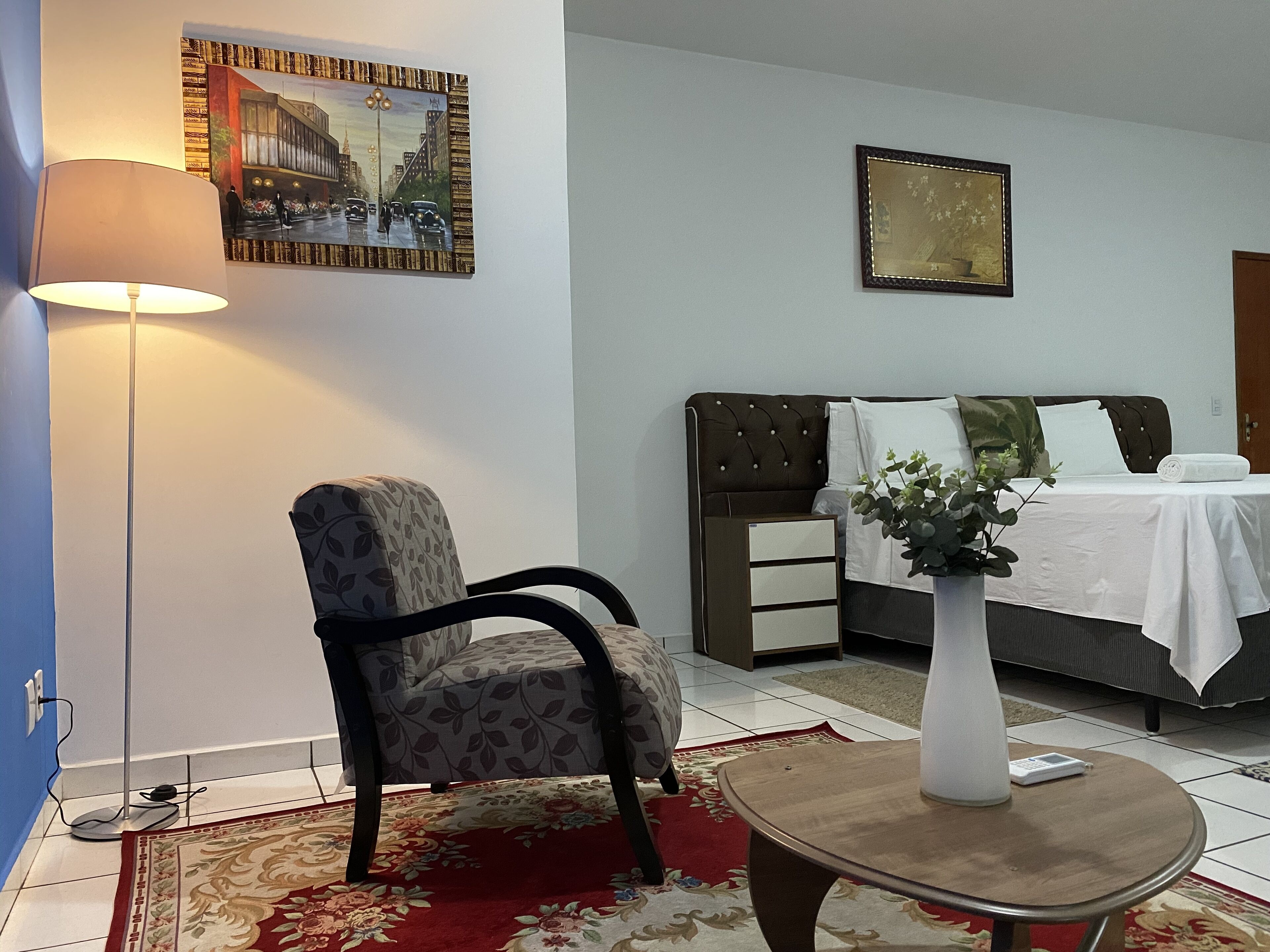 A hotel room with a bed, a chair, a lamp, a vase with flowers, and a painting on the wall.