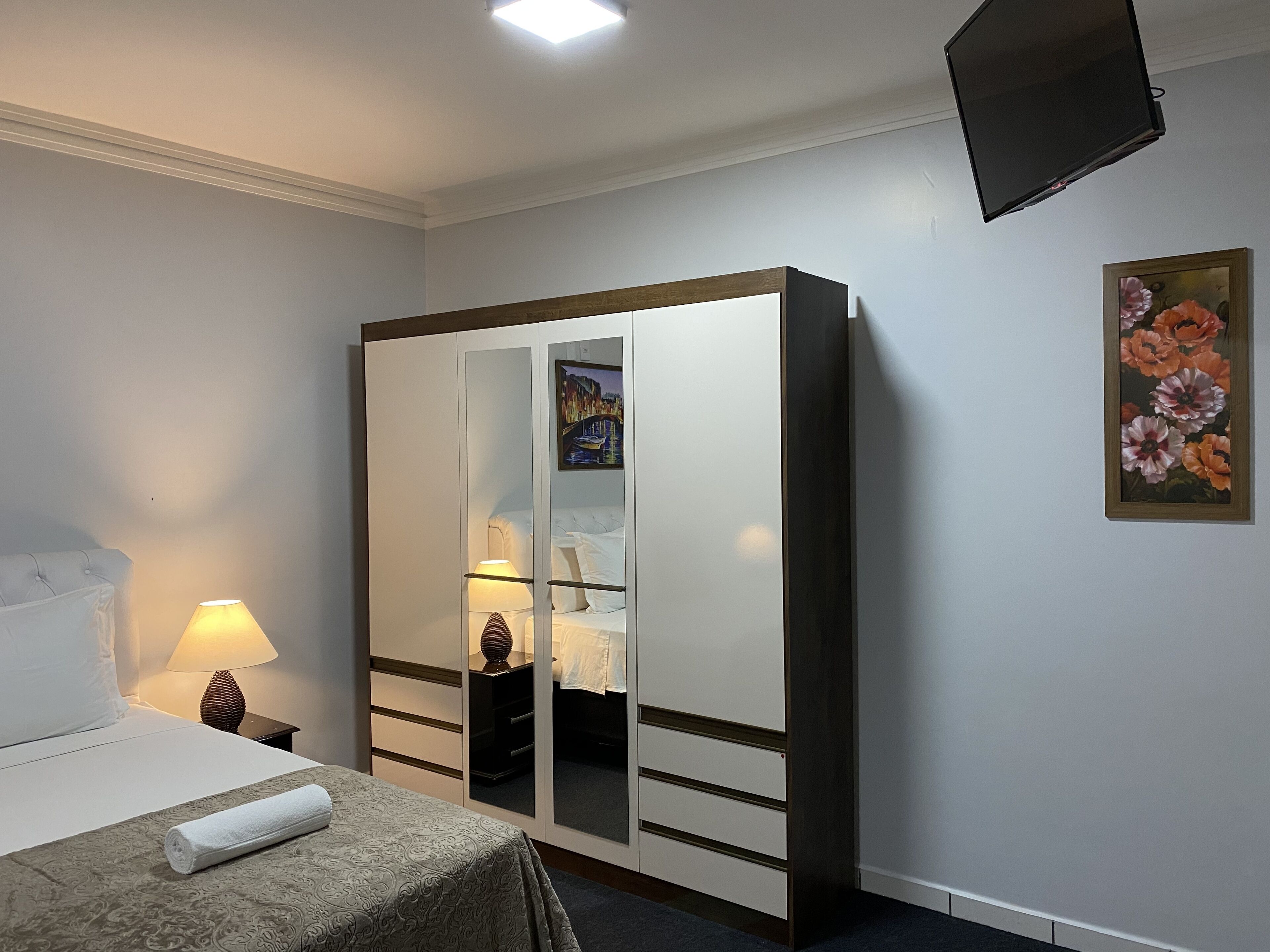A hotel room with a bed, a wardrobe with a mirror, a lamp, and a painting on the wall.