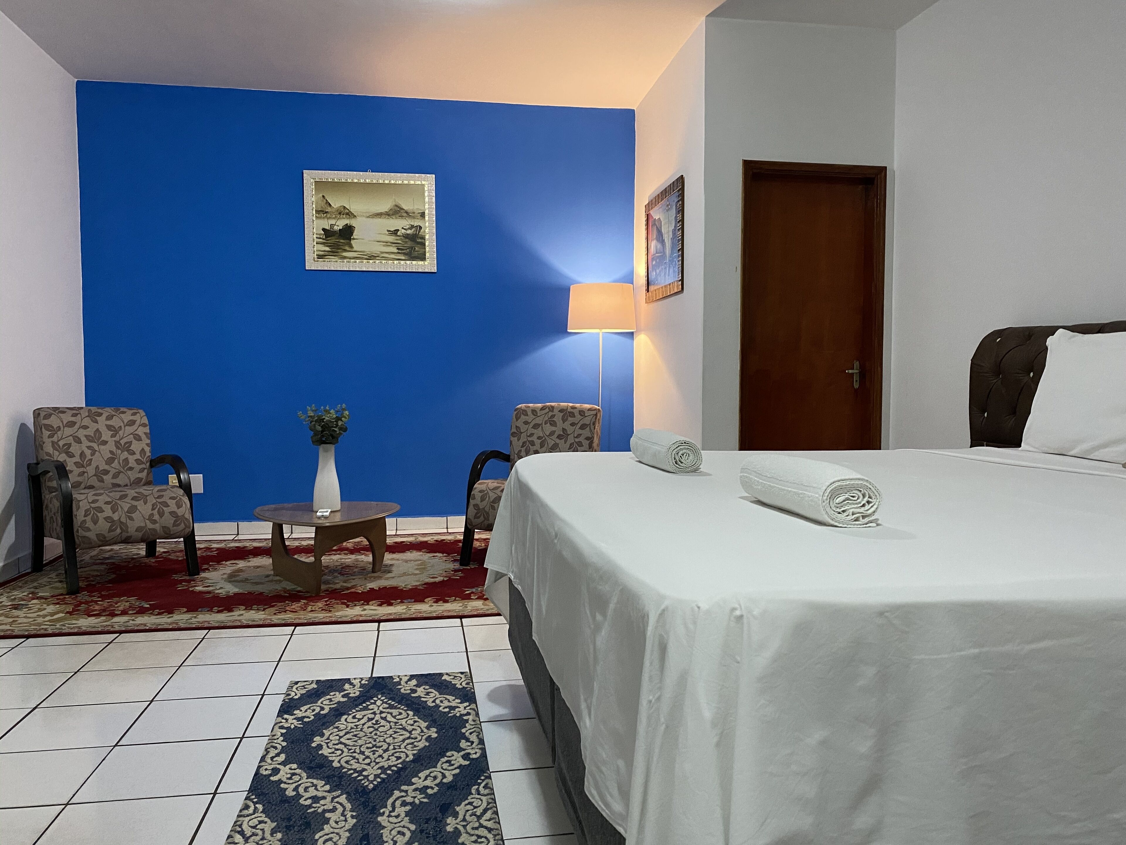 A hotel room with a blue accent wall, a bed with white linens, two armchairs, a small table with a vase, and a lamp.