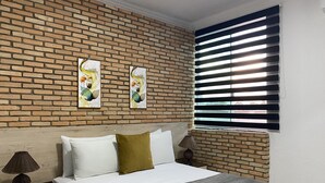Ein Schlafzimmer mit Backsteinwand, einem Bett mit weißen und grünen Kissen, einem Nachttisch mit Lampe und einem Fenster mit Jalousien.