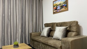 Ein Wohnzimmer mit einer Couch, einem kleinen Holztisch mit Pflanze, grauen Vorhängen und einem Bild an der Wand.