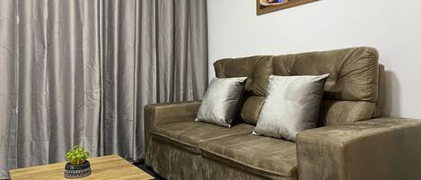 A living room with a sofa, a small wooden table with a plant, grey curtains, and a painting on the wall.