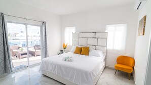 Ein modernes Hotelzimmer mit einem großen Bett, einem leuchtend orangefarbenen Sessel und Blick auf die Stadt durch eine Glastür.