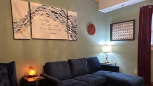 Ein Wohnzimmer mit einem dunkelblauen Sofa, einem kleinen Tisch mit einer Lampe, einem roten Dekorationsplatten und gerahmten Kunstwerken an der Wand.