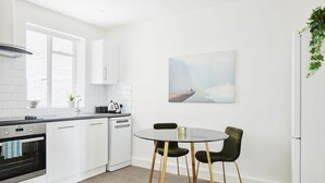Une cuisine moderne avec des meubles blancs, un coin repas comprenant une table ronde et deux chaises, et une grande fenêtre laissant entrer la lumière naturelle.